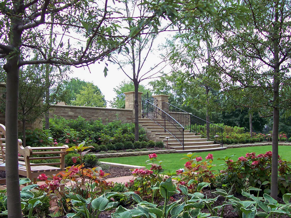Garden view through trees with lawn, stone walls, steps, bench, flowering beds, and terrace railings.